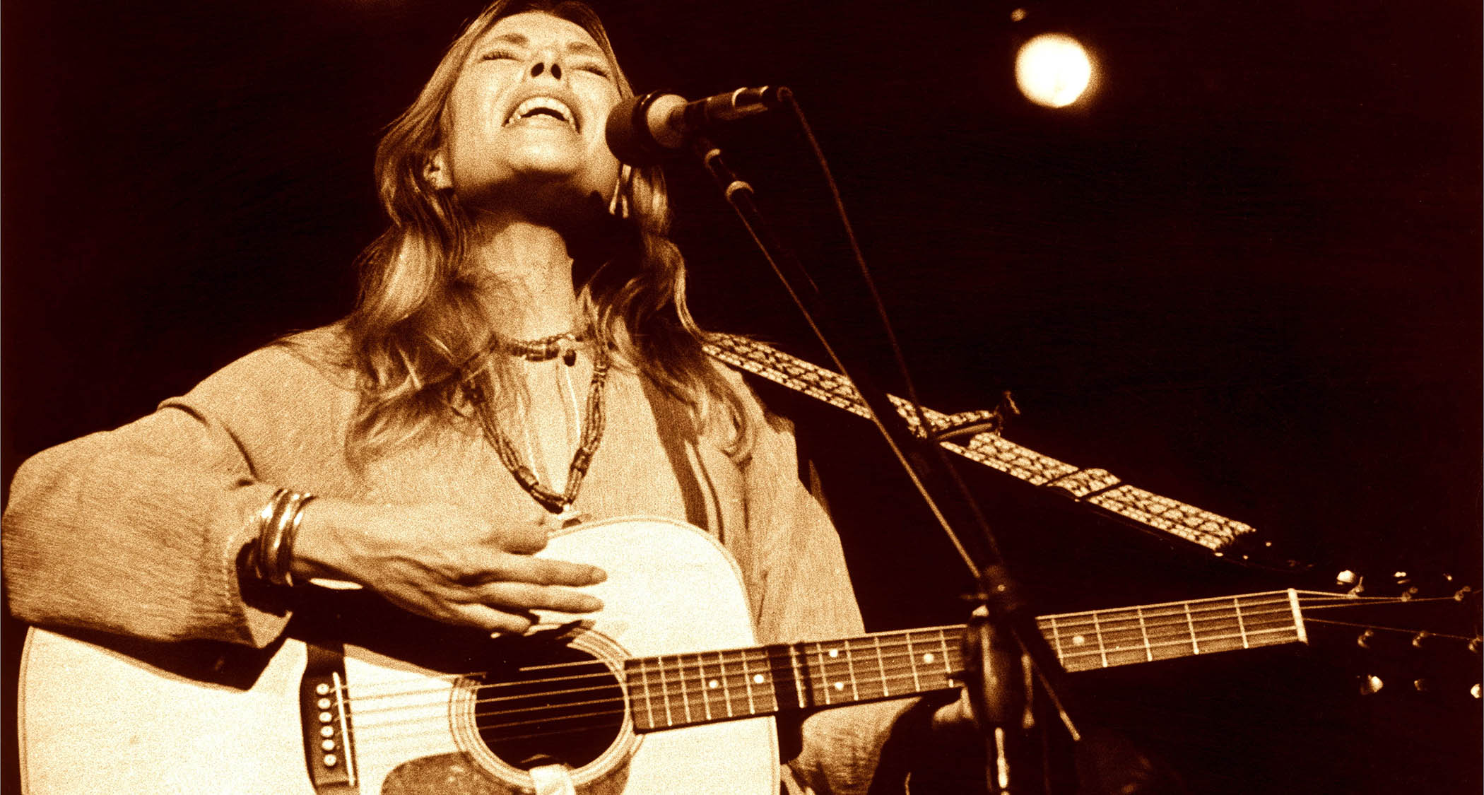 A classic sepia-toned live shot of Joni Mitchell playing her Martin dreadnought and singing live in 1974