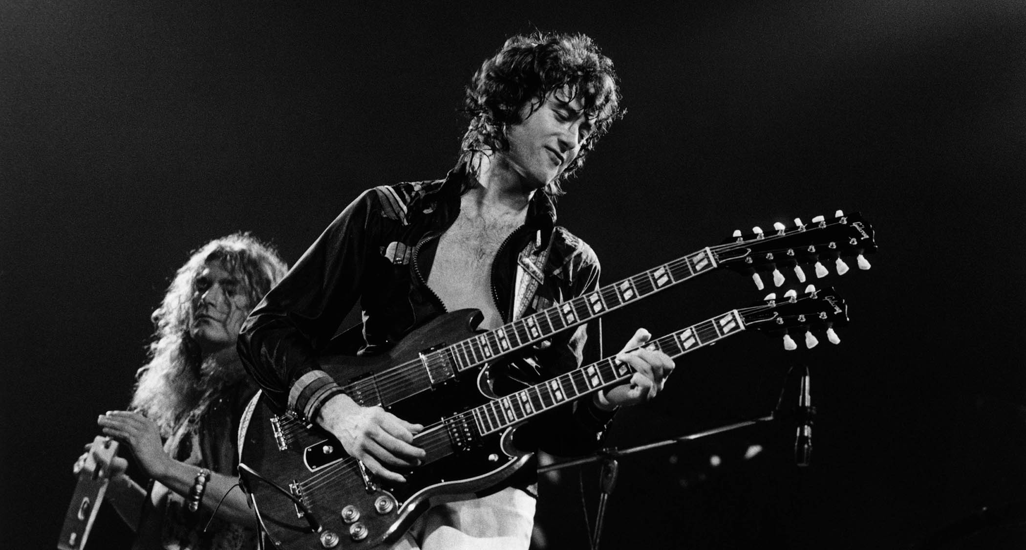 Jimmy Page plays his double-neck onstage with Led Zeppelin as frontman Robert Plant plays the tambourine in the background.