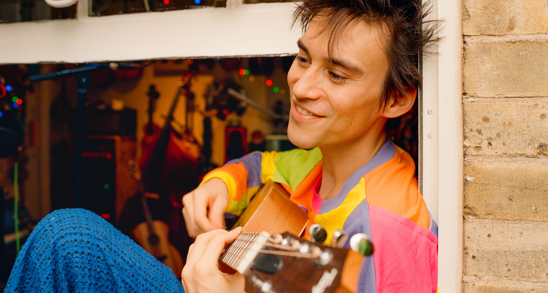 Jacob Collier sits in a windowsill and plays an acoustic guitar. he wears a multi-colored shirt.